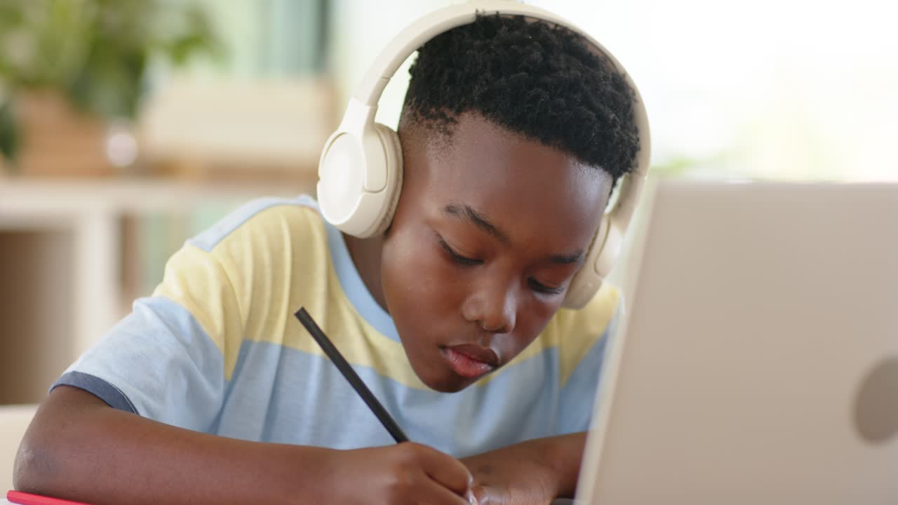 African American boy studying at home desk writing in notebook with headphones and checking laptop