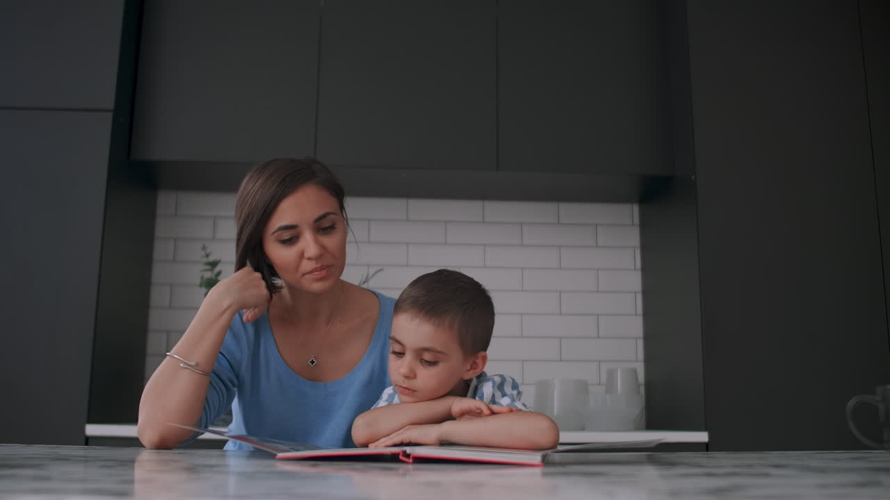 joven madre y hijo españoles sentados a la mesa leyendo un libro juntos y sonriendo