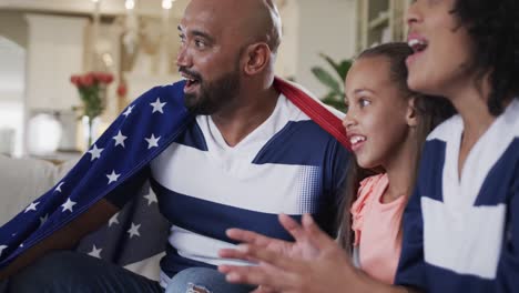 padres biraciales emocionados e hija viendo deportes en la televisión con bandera estadounidense en casa, cámara lenta