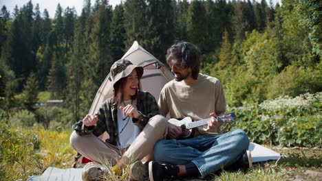 A-guy-and-a-girl.-Tourists-on-a-fun-vacation-sit-near-the-tent,-play-the-guitar-and-sing-songs-against-the-backdrop-of-a-green-forest