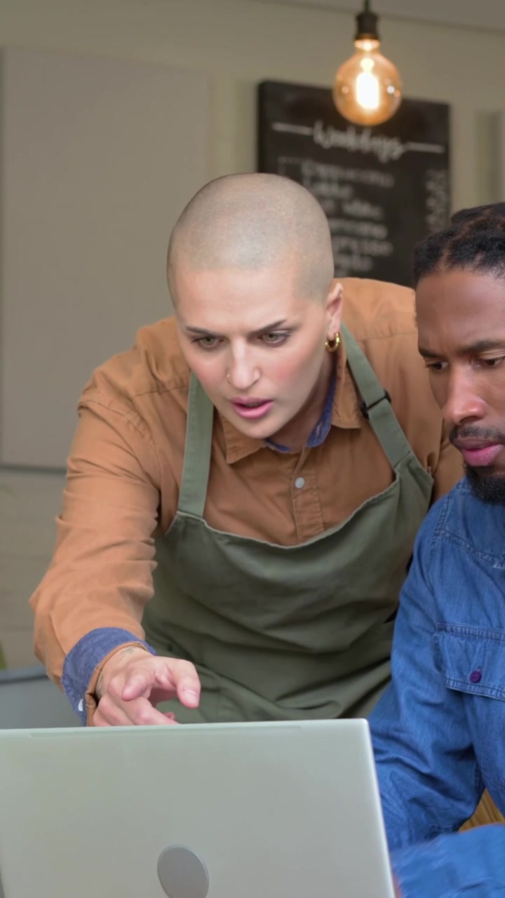 Vertical video: Seeing guy gesturing staff in green apron walking to seat helping on laptop in cafe