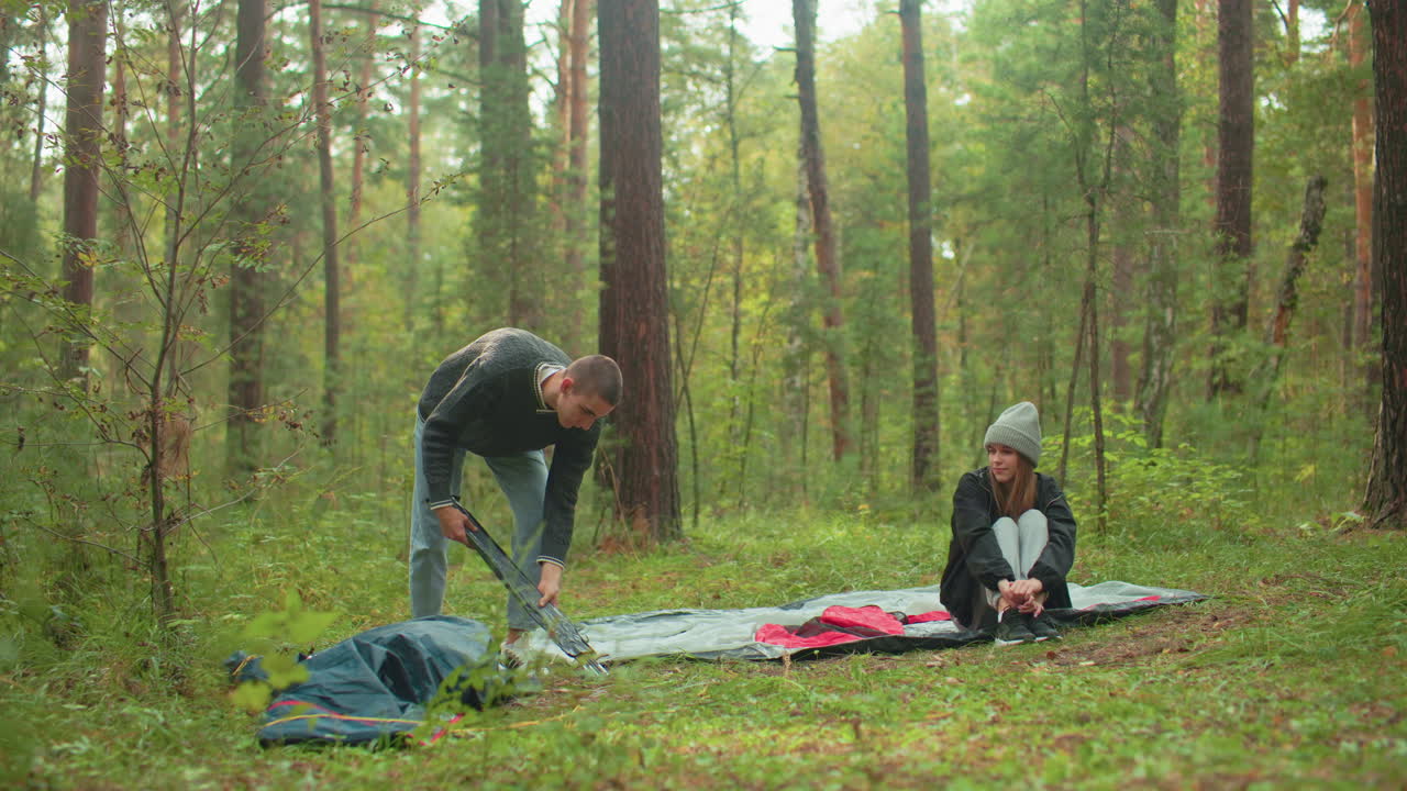 Boy in sweater unpacks tent poles while standing beside scattered gear in forest, girl sits cross-legged on tent fabric wearing jacket and beanie, both preparing for camping setup surrounded by trees