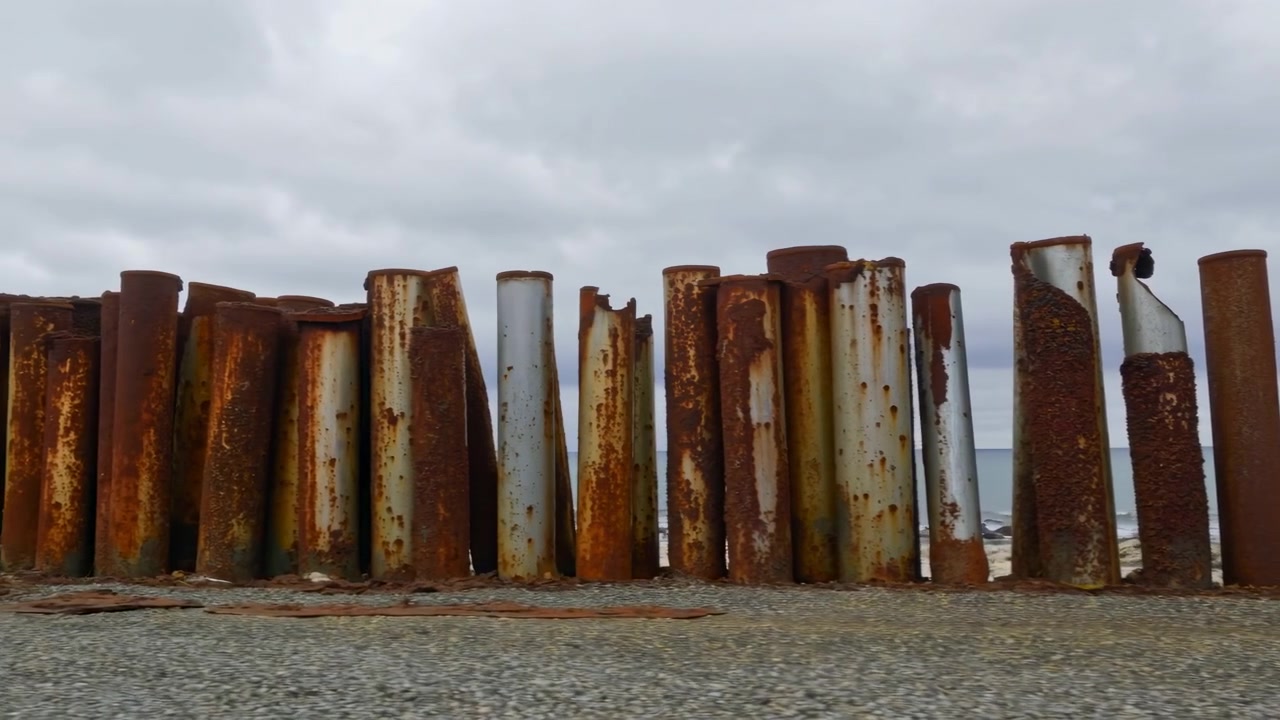Høje cylindriske stålpæle står på række langs kystlinjen, stærkt korroderet af saltvand med orangebrun rust, afskallende skorper, grubet tekstur og blødt gråt kystlys, med rust, pæl