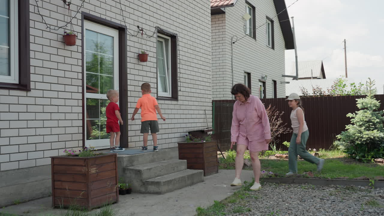 Children Enter Suburban Home, Mother Watches. Brick Facade And Glass Door Open Onto Concrete Steps And Wooden Planter Beside Fenced Lawn. Woman In Pink Robe Guides Two Children Into House With Gentle