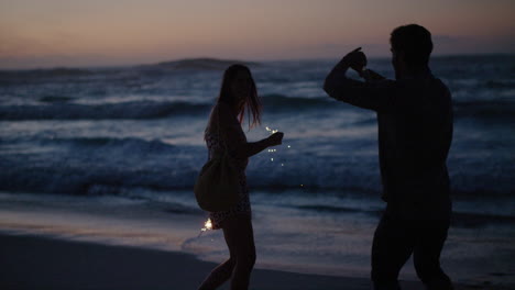 young man taking photo of girlfriend holding sparklers dancing celebrating new years eve laughing playful together on beach at sunset slow motion