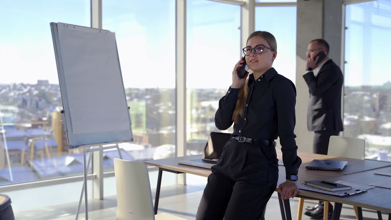 Beautiful secretary and her boss in office. Smiling young business woman holding a phone on the background of a businessman and talking the phone