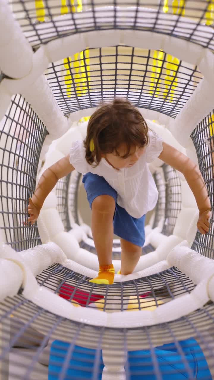 A happy 5-year-old girl crawls through a soft, netted play tunnel with a smile in slow motion, having fun exploring an indoor playground - vertical slow motion
