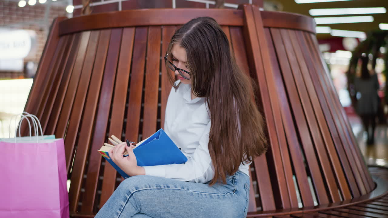 cerca de transeúntes ocupados con una dama sentada, piernas cruzadas, profundamente involucrada en la lectura de un libro, bolsas de compras a su lado en un banco de madera en un bullicioso centro comercial