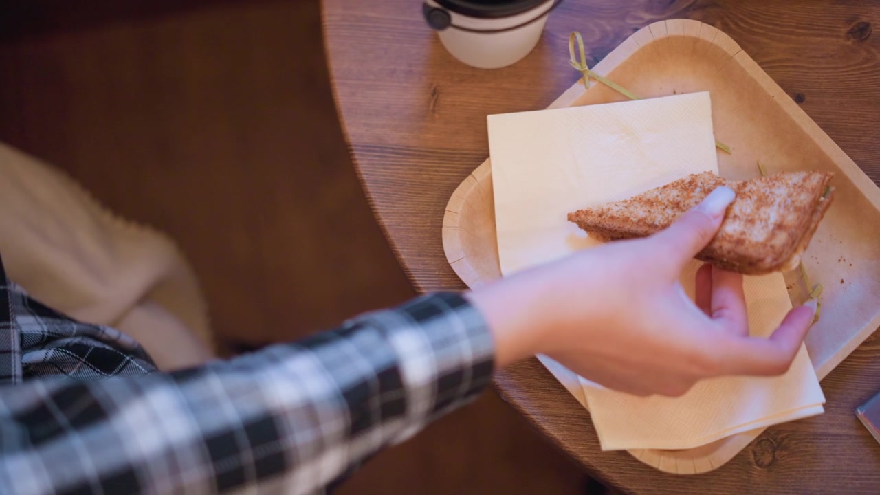 Young woman in plaid shirt reaches for and picks up toasted sandwich from wooden tray on table with napkins visible. scene is set indoors at dining area with warm lighting showing woman arm