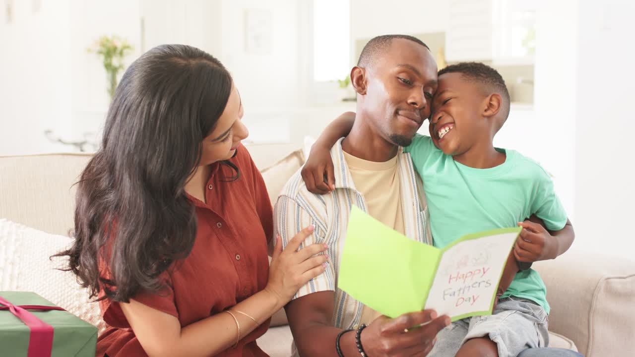 Diverse family surrounding father reading green Father's Day card, celebrating and hugging on couch