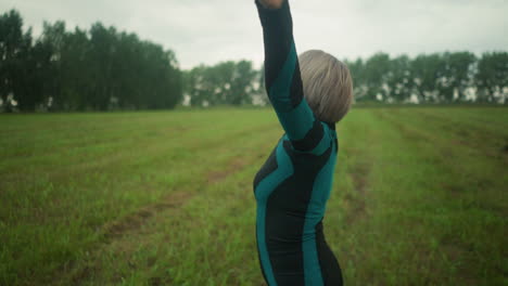 side view of middle-aged woman outdoors in a vast grassy field practicing yoga, she is slowly lifting and bringing down her hand, focused on her movements