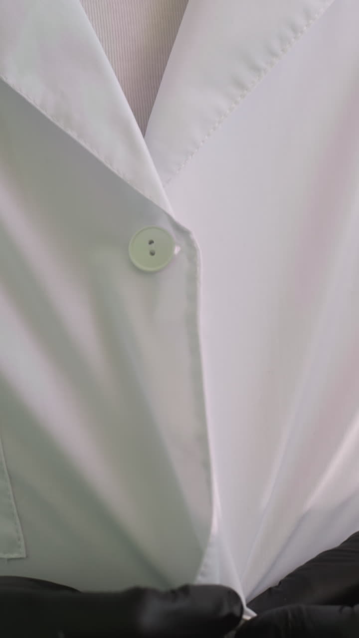 A close-up view of someone wearing black gloves as he carefully buttons his lab coat, with a view of a shelf behind