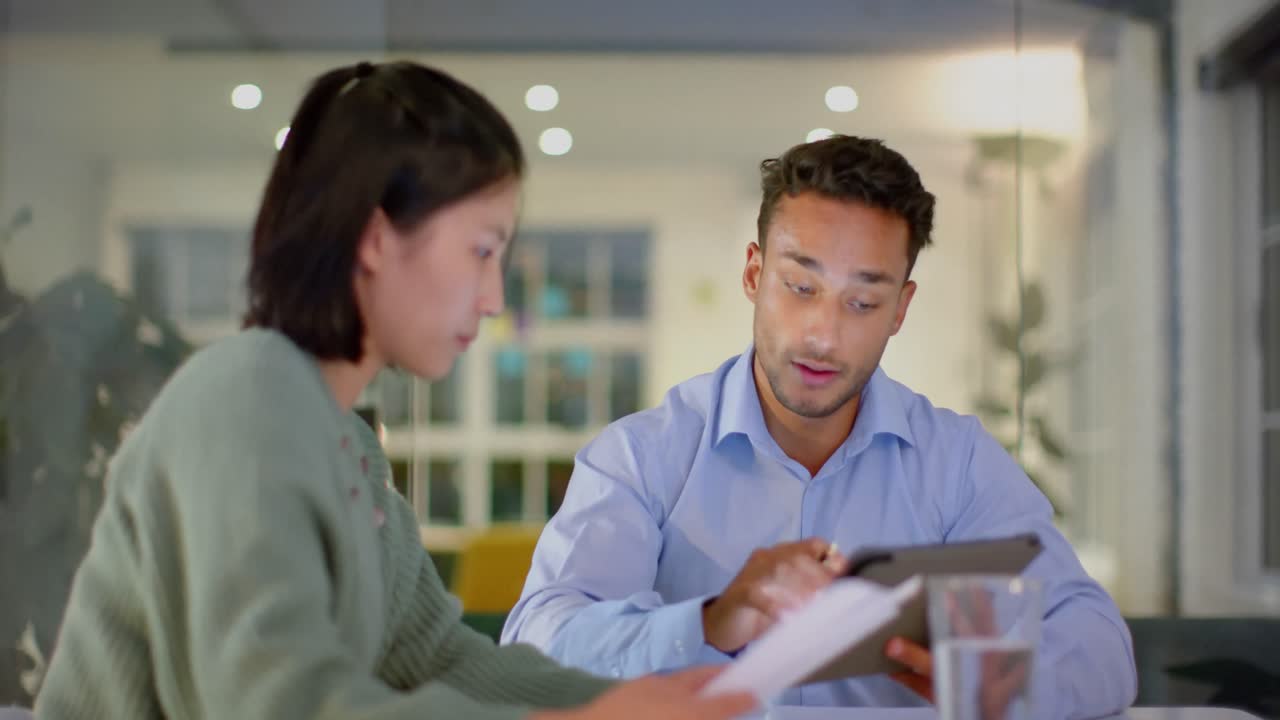 Diverse male and female colleague discussing work and using tablet late at office
