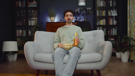 young man watching interesting movie on tv while eating chips and drink soda sitting on couch at home