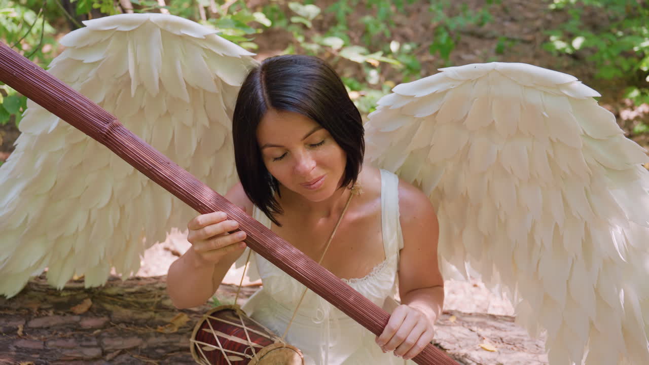 Overhead view of forest guardian with white wings seated on woodland ground, gently holding wooden staff near chest, bathed in warm sunlight, expressing serenity
