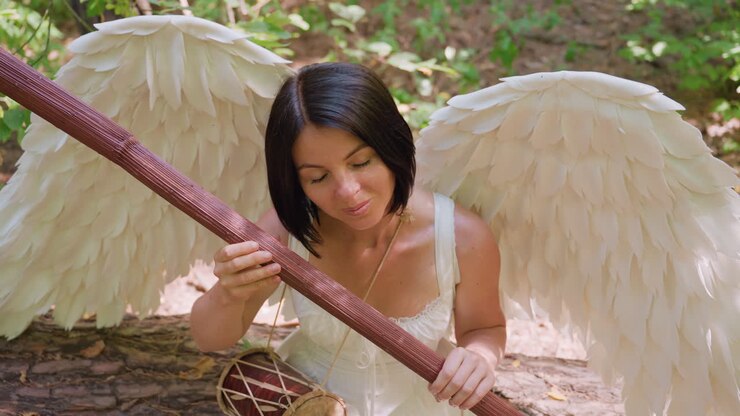 Overhead view of forest guardian with white wings seated on woodland ground, gently holding wooden staff near chest, bathed in warm sunlight, expressing serenity