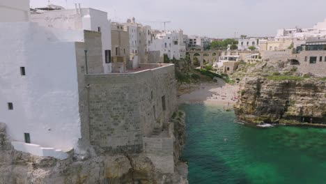 Slow-aerial-footage-reveal-of-cliffside-buildings-and-then-a-cove-with-a-beach-in-Polignano-a-Mare,-Italy