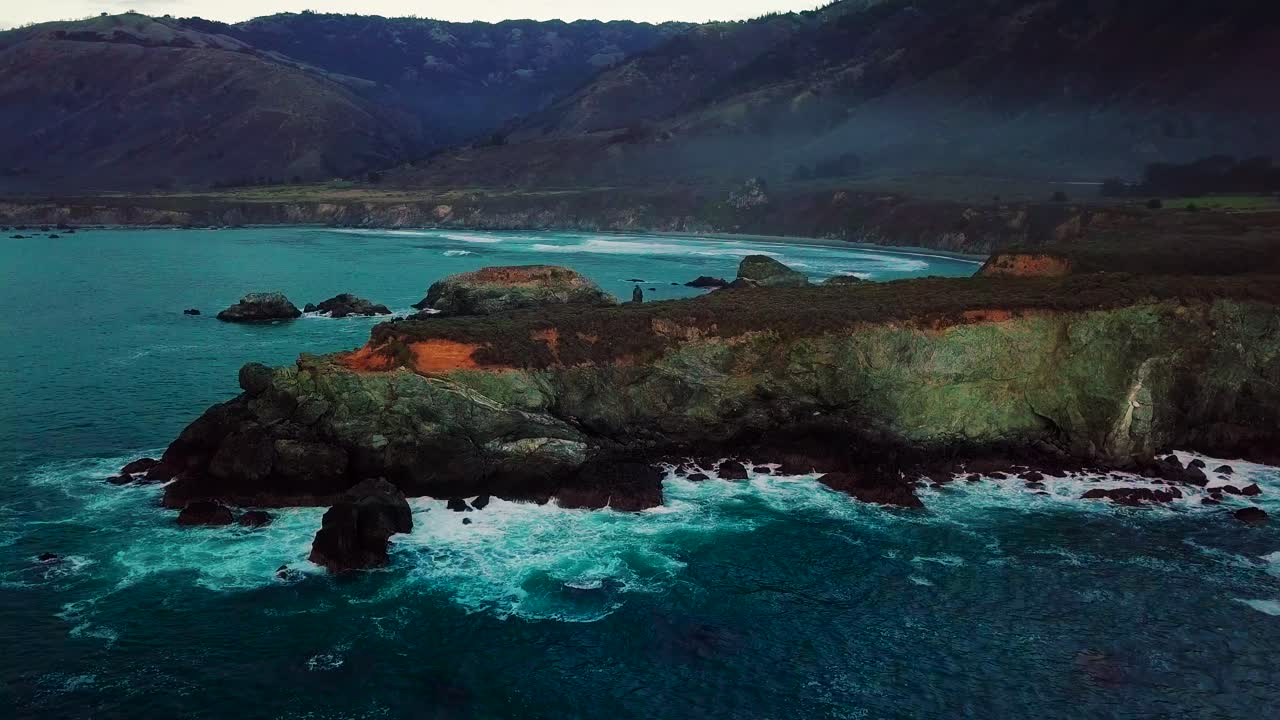 Static aerial wide view of waves crashing on rocky ocean cliffs at Sand Dollar Beach in Big Sur California at twilight