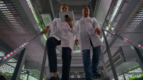 Men-in-white-lab-coats-tablet-computer-down-the-stairs-of-a-modern-vertical-farm-camera-moves-behind-them-and-shows-2-women-in-white-lab-coats-and-glasses-discussing-plant-samples