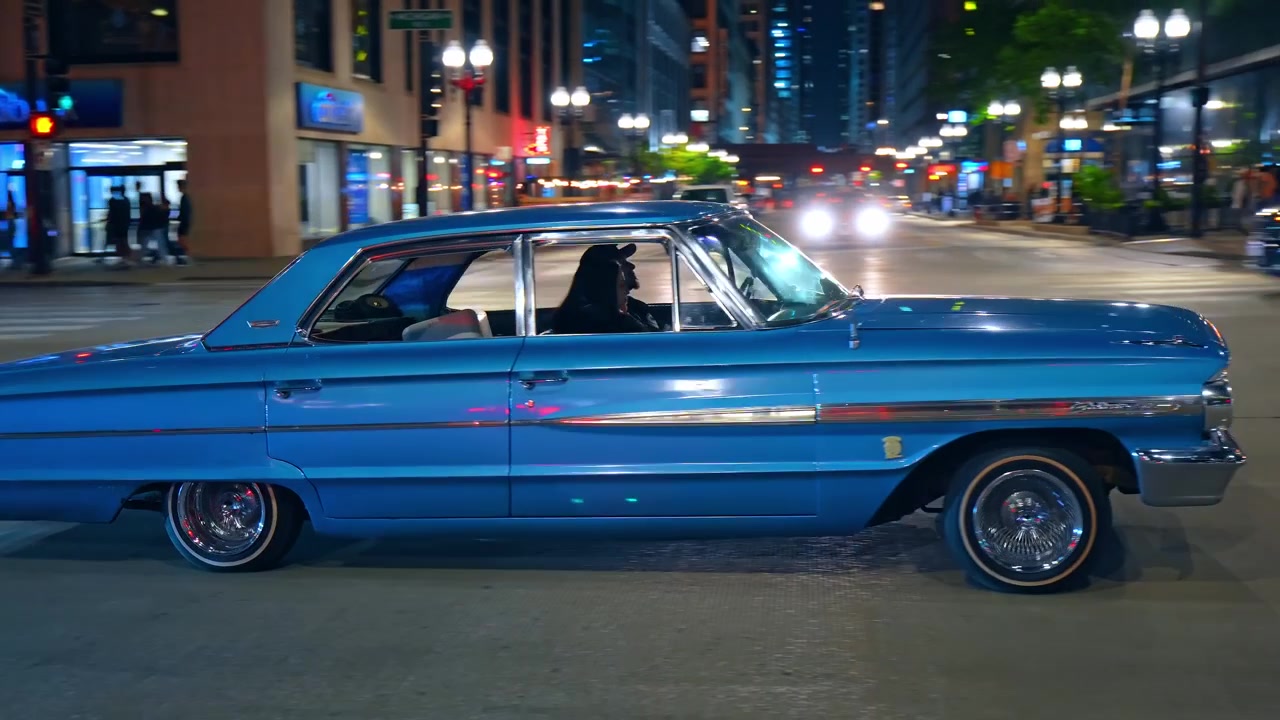 Chicago, USA, 7. Mai 2026: Klassischer blauer Lowrider fährt nachts durch eine Innenstadtstraße in Chicago. Seitenansicht mit Chromspeichenfelgen und Neon-Schaufenstern im Hintergrund.