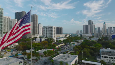 Fliegen-Sie-Um-Die-Amerikanische-Nationalflagge-Herum,-Die-Auf-Der-Stange-über-Der-Stadt-Weht.-Büro--Oder-Wohnhochhäuser-Im-Hintergrund.-Miami,-USA