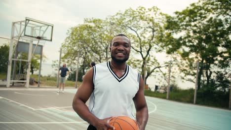 Portrait-of-a-young-Black-man-who-is-posing-looking-at-the-camera-and-smiling-in-front-of-his-friends-who-are-playing-basketball-outside-in-the-summer