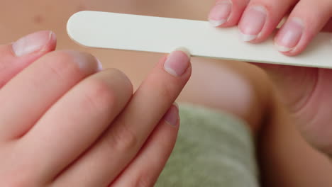 close up hand view of lady filing fingernails using white nail file while wrapped in towel, showing personal hygiene and beauty care activity in indoor setting focused on natural grooming routine