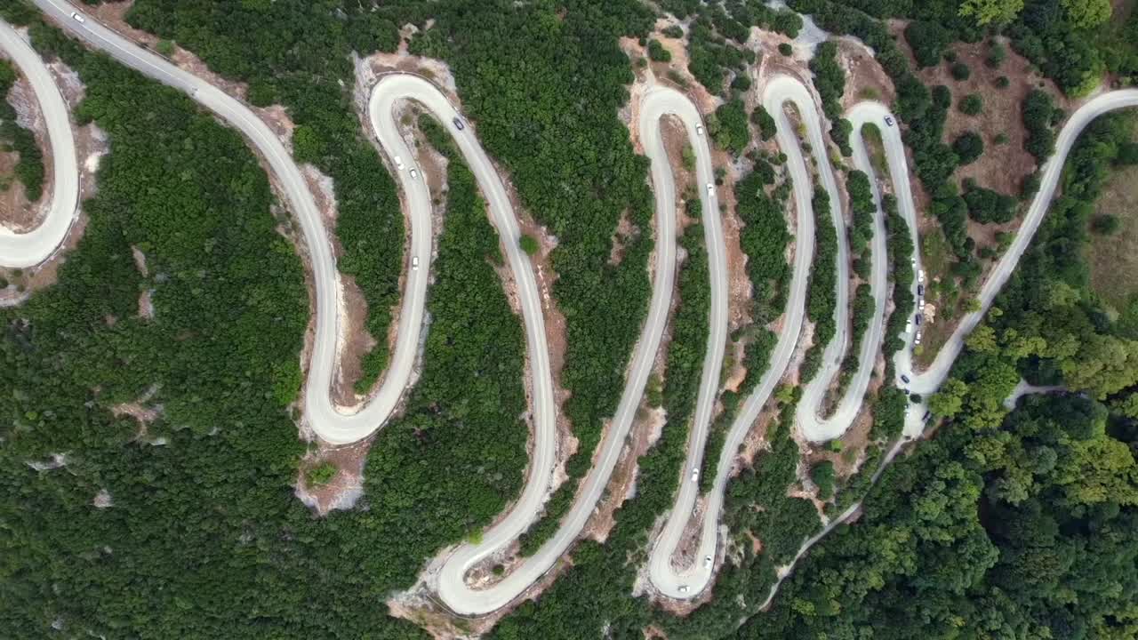 Cinematic top-down drone ascending shot of the famous zig-zag road in Papigo, Greece. High-angle view of white cars navigating the sharp hairpin turns through a lush forest. Ideal for travel films