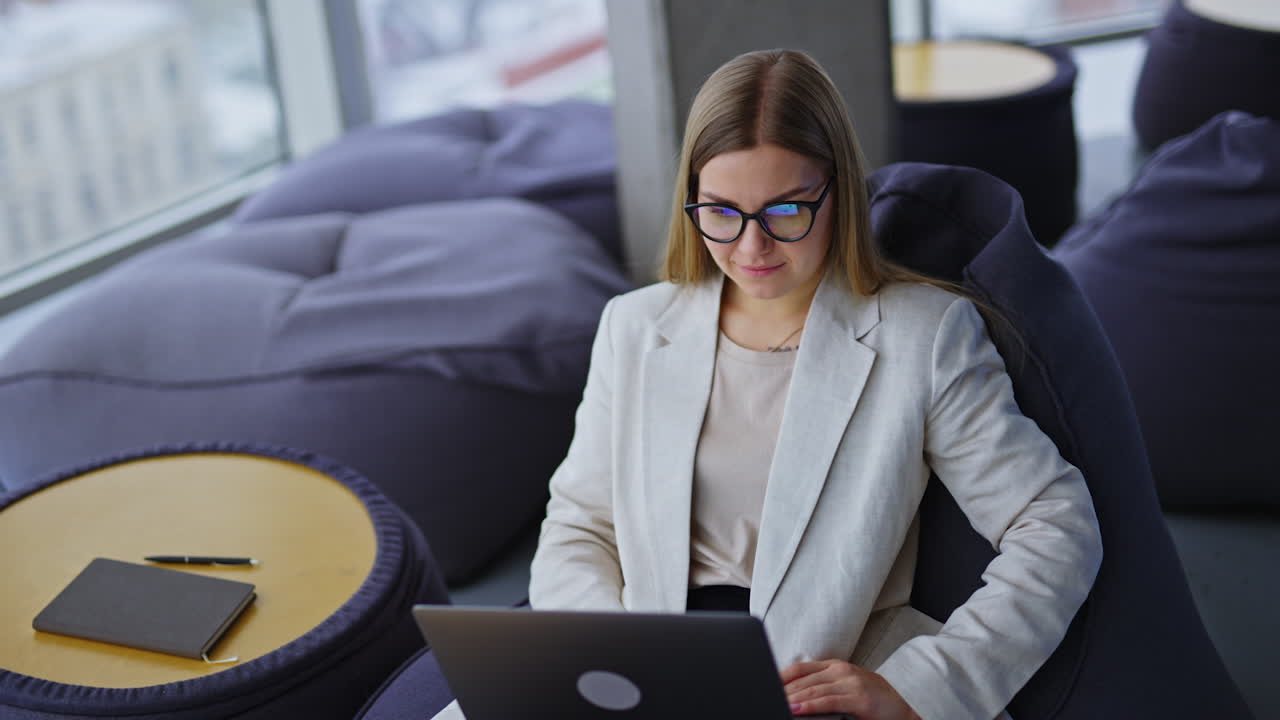 Lady in light jacket sitting in bean bag chair working on her laptop. Comfortable workplace in a nice modern creative office. Blurred backdrop.