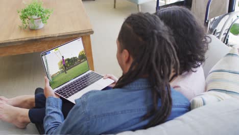 Video-Von-Zwei-Afroamerikanern,-Die-Auf-Der-Couch-Sitzen-Und-Sich-Ein-Fußballspiel-Auf-Dem-Laptop-Ansehen