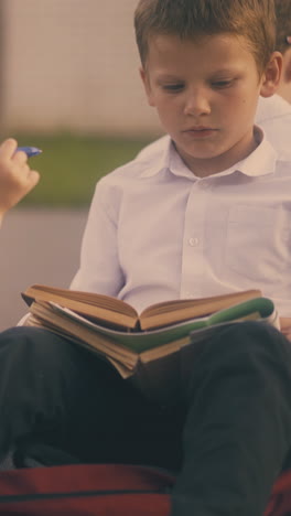 funny little girl with ponytail holds blue pencil talks to schoolboy reading textbook with friend in sunny street close