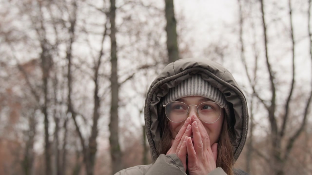 Mujer caucásica mirando hacia arriba en un parque, vistiendo abrigo con capucha y gafas, manos entrelazadas cerca del pecho en gesto de agradecimiento, árboles desnudos y luz otoñal tenue, reflejo sereno y expresión esperanzadora capturados