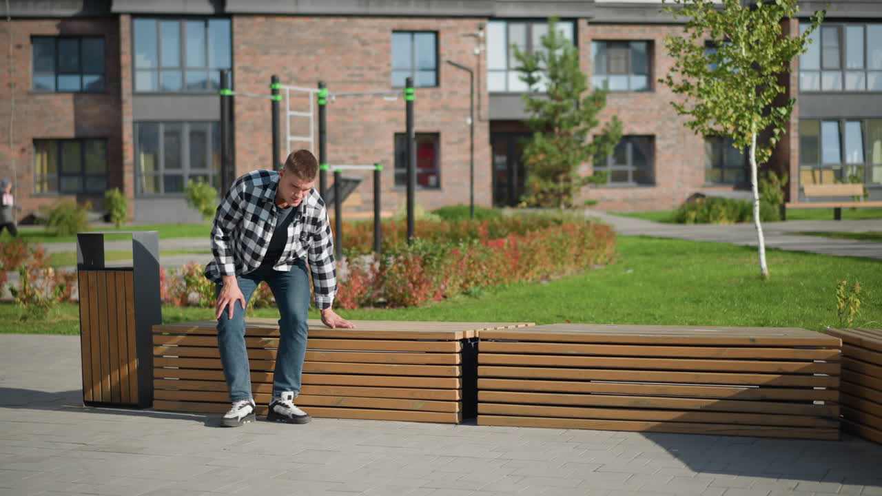 young man in checkered shirt walks to wooden bench and sits down to retrieve medication from pocket while another person walks near brick building in soft blur background under sunny weather