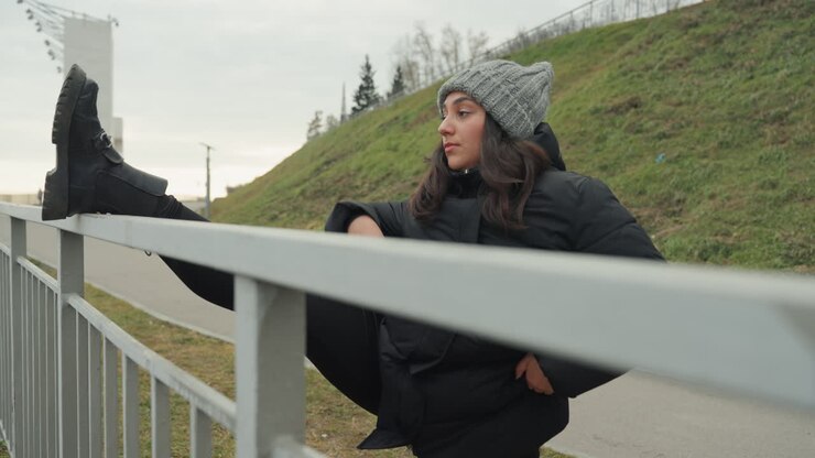 Ballerina latina su una ringhiera che allunga lo stivale e si equilibra in cappotto invernale, berretto e stivali pesanti, su sfondo di collina e ponte all'aperto, momento di riscaldamento spontaneo che mostra movimento, sguardo e flessibilità controllata.