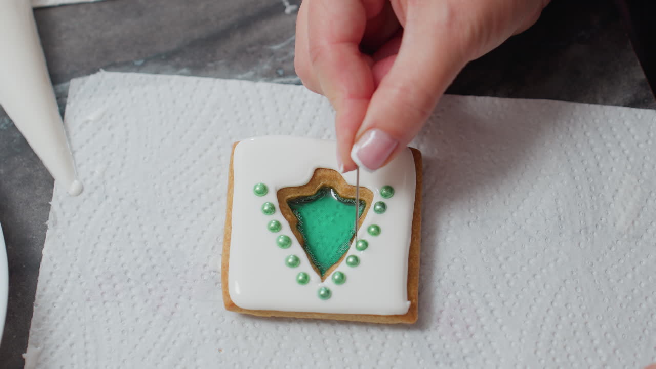 Close-up of hands adjusting green decorative beads on iced cookie with white icing and green filling, the cookie is placed on a tissue paper with intricate details of bead placement for decoration