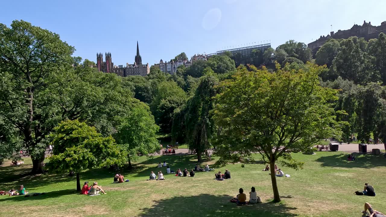 Wide-angle daytime footage of a green city park in Edinburgh, Scotland, showing people lounging on grass under clear skies, with slow camera pan revealing historic skyline