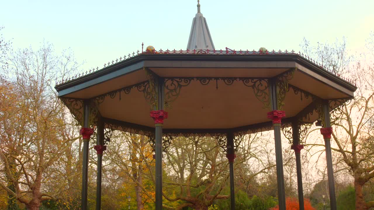 Billede af den historiske, victorianske musikpavillon i Battersea Park, London, taget i lav engel. Fremhæver det indviklede jernarbejde og de røde detaljer mod en baggrund af efterårstræer og en blød, gylden himmel.