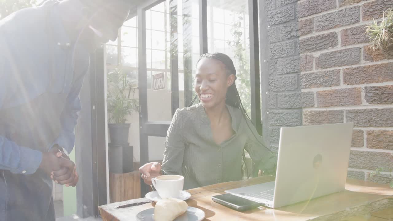 African american male barista serving female customer using laptop outside coffee shop, slow motion