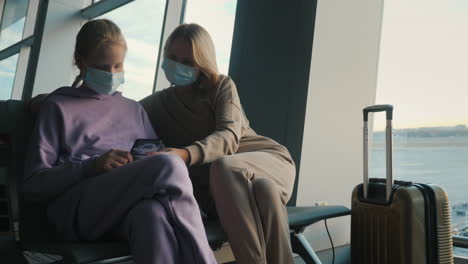 mother and daughter waiting for their flight in the airport terminal. they are wearing protective masks, using a smartphone