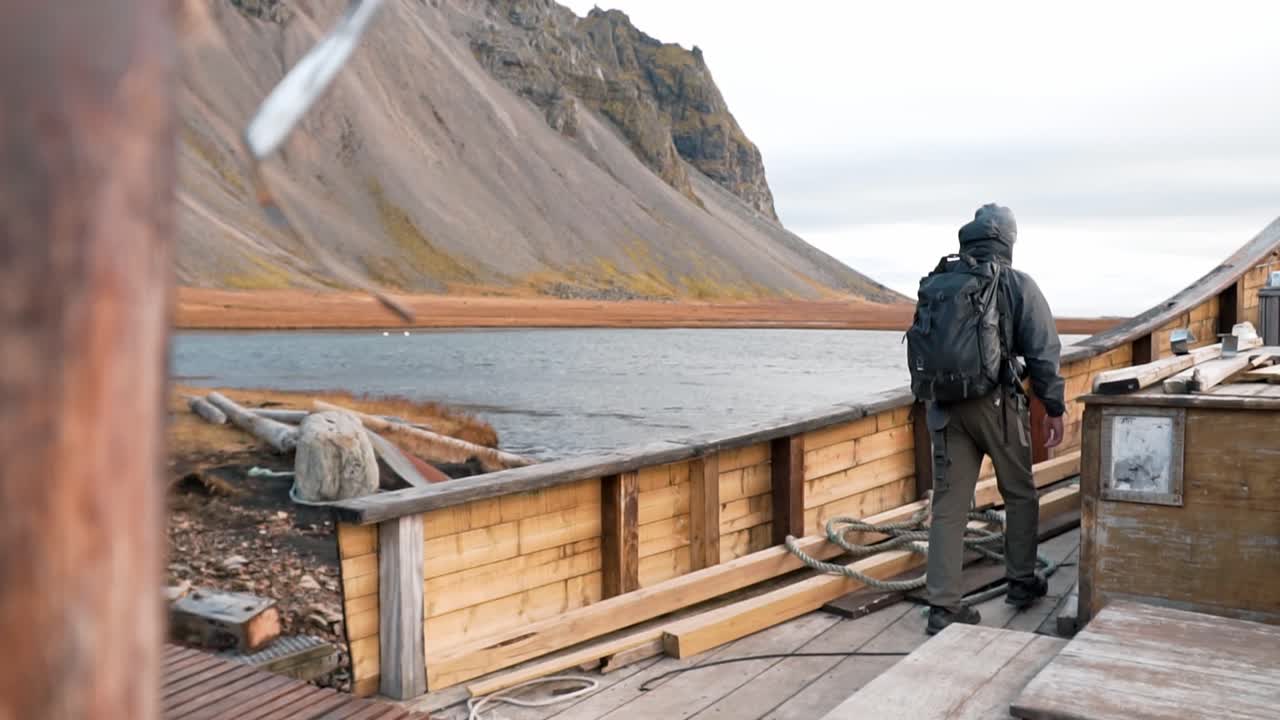 Traveler with a backpack walks on a Viking boat, gazing out at the serene waters with the iconic Vestrahorn mountains dominating the background at Stokksnes, Iceland