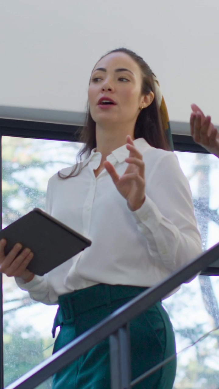 Vídeo vertical: Mulher de blusa branca descendo a escada, segurando um tablet e gesticulando, enquanto um colega de equipe se aproxima.