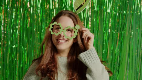 woman celebrating st patrick's day standing in front of green tinsel curtain wearing prop shamrock shaped glasses