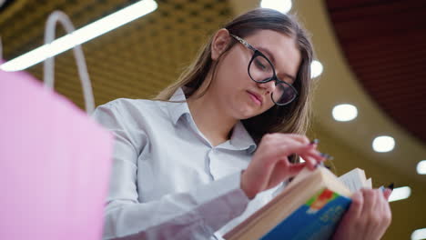 primer plano de una mujer pensativa con gafas, leyendo atentamente un libro, con una bolsa de compras rosa borrosa en el fondo, en un animado centro comercial