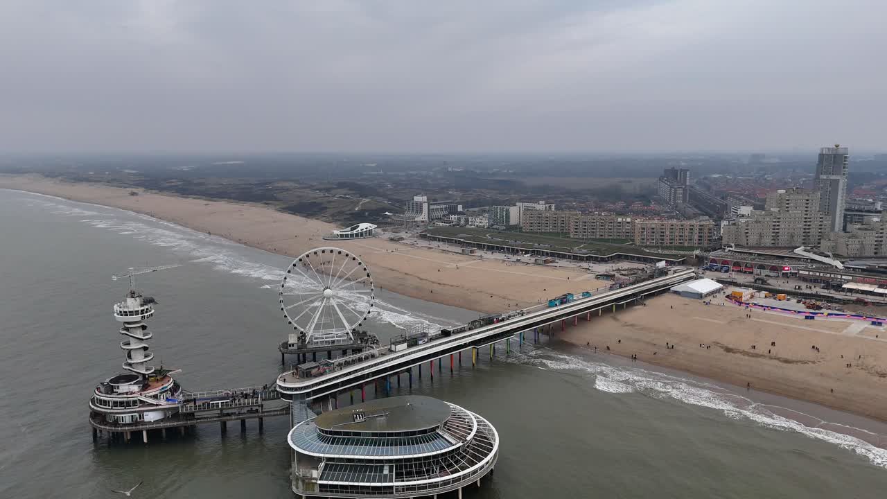 Ferris Wheel The Pier At Scheveningen City, The Hague, Netherlands During cloudy day in winter. Sandy beach and walking tourist on bridge. Apartment housing blocks in distance. Aerial wide shot.
