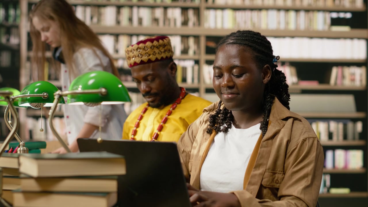 Black woman student uses laptop at shared library table with stacked books