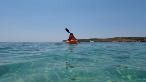 Vista-Del-Nivel-Del-Agua-De-Una-Mujer-Con-Un-Kayak-Naranja-Que-Pasa-Por-El-Mar