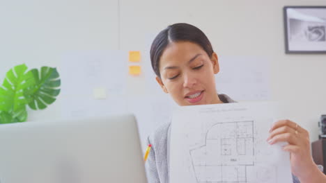 female architect with laptop in office sitting at desk showing plans for new building on video call
