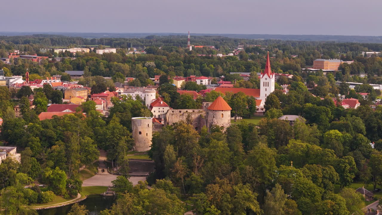 Aerial establishing left of Cesis, Latvia, showcasing a historic town center surrounded by lush greenery and slight haze in air