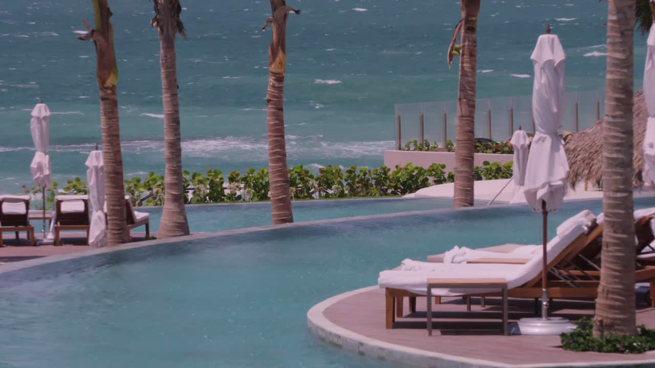 A wide shot on a windy day captures an infinity pool with lounge chairs and palm trees, overlooking a turbulent ocean with crashing waves at a tropical resort.