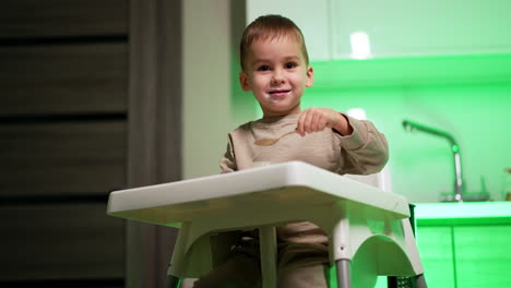 adorable caucasian toddler sitting at the high chair eating dairy. kid uses a spoon, talks and smiles to camera. low angle view.